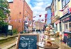 A lively street scene in a historic town, featuring colorful buildings, cafés, and people walking along the cobblestone path.