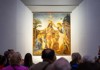 A group of travelers listens intently to a guide in front of a golden religious painting displayed in a softly lit museum hall.