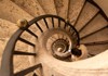 A spiral staircase with ornate stone steps winds downward, with a visitor making their way to the lower level.