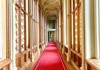 An ornate hallway inside a royal residence features tall windows, golden trim, and a red-carpeted walkway.