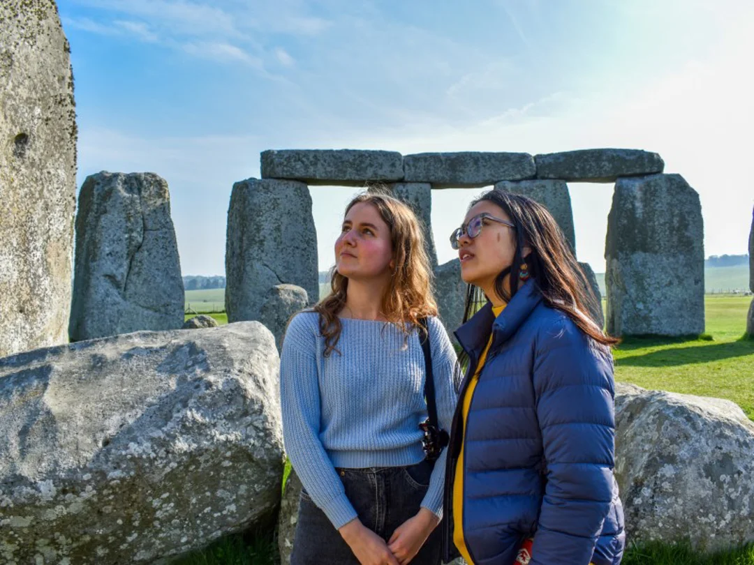 Two women pose in front of Stonehenge, smiling as they stand close together with the ancient stones behind them.