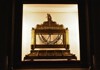 A glass-encased religious relic displayed in a dimly lit chamber, framed by golden decorations.