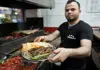 Grill cook presenting a plate of grilled meat, flatbread, and peppers inside a kebab shop
