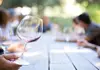 Person holding a partially filled wine glass at an outdoor tasting table with blurred group in background.