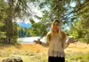 Woman standing beneath pine trees near an alpine lake with mountains in the background