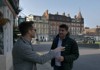Two men shake hands near a grand historic building, engaging in conversation with an elegant cityscape behind them.