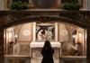 A visitor stands inside a small, dimly lit chapel, gazing at an illuminated religious painting framed by classical architecture.
