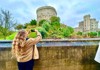 A visitor in a cozy brown coat takes a photo of a medieval castle, standing by a peaceful river lined with stone walls.