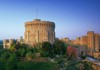 Windsor Castle stands tall atop a lush green hill, bathed in golden sunlight with a clear blue sky in the background.