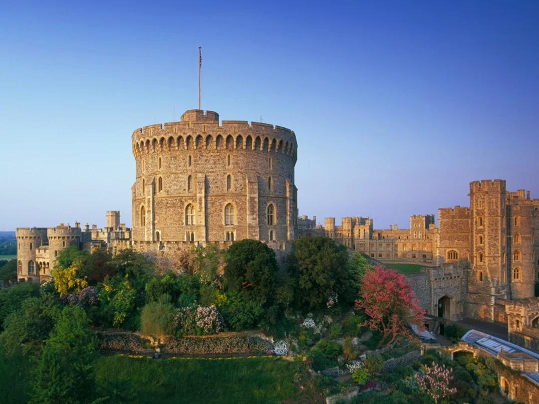 Windsor Castle stands tall atop a lush green hill, bathed in golden sunlight with a clear blue sky in the background.