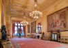 A lavish royal hall adorned with intricate chandeliers, golden-framed paintings, and plush furniture exudes opulence.