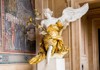 A gilded angel statue with flowing ribbons and outstretched wings stands as an ornate decoration inside a sacred space.