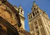 La Giralda bell tower and nearby cathedral structures in Seville, shown from a low angle against a blue sky.