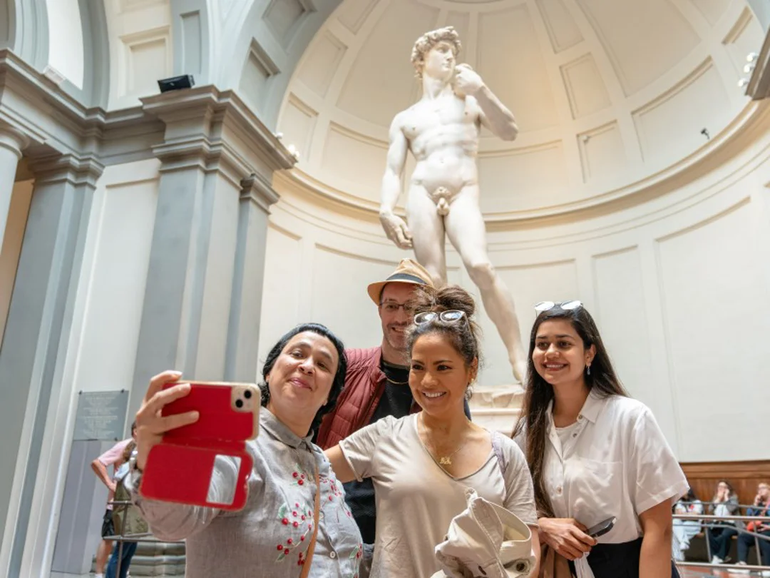 A tour guide enthusiastically gestures while speaking to two visitors in front of Michelangelo’s David statue inside the Accademia Gallery.