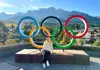 Woman seated in front of the Olympic rings with a mountain village and peaks behind her