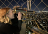 A woman standing on the summit of the Eiffel Tower looking through a telescope at night.
