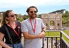 A tour guide speaks to a woman near the Roman Forum, with lush greenery and ancient ruins in the background.
