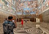 A tour guide gestures toward the ornate ceiling inside the Sistine Chapel, explaining the artwork to a small group of tourists.