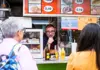 Street food vendor speaking to customers at a snack stand displaying menus and drinks in Vienna.