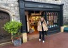 A woman in a stylish winter outfit stands in front of a quaint bookstore, smiling as she prepares to step inside.