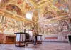 A group of tourists poses inside a Vatican Museum room, surrounded by vibrant murals and historical artifacts.