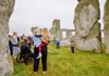 A tour guide gestures while explaining the history of Stonehenge to a small group of visitors standing among the ancient stones.