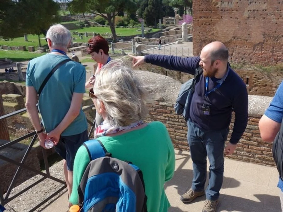 Meet your guide at Cafè Piramide Guide pointing to the ruins of Ostia Antica with a group.