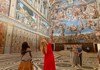 A woman walks through a vast hall in the Vatican Museums, gazing up at the elaborate frescoes covering the ceiling.