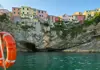 Rocky coastal cliffs of Cinque Terre with pastel houses above, seen from a boat with a lifebuoy in the foreground.