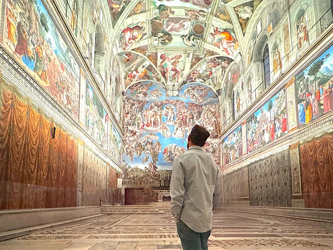 Visitors walk through the Sistine Chapel, admiring the intricate frescoed ceilings illuminated by warm light.