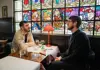 Two people sitting at a restaurant table with stained-glass windows behind them, talking over drinks and water.