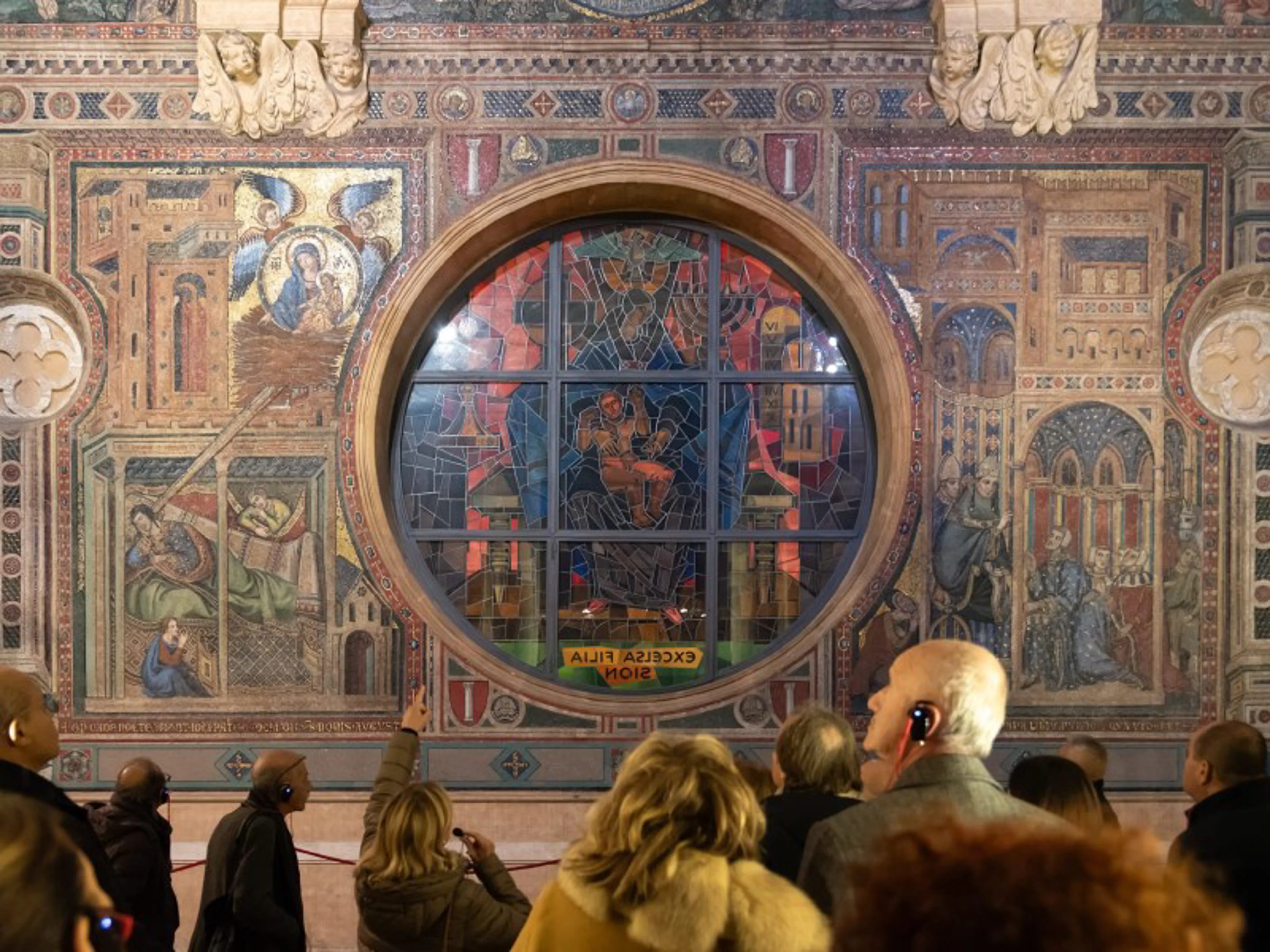 Visitors admire an intricate mosaic inside a historic church, with golden accents and religious iconography decorating the walls.