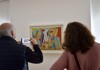 Two visitors engage in discussion while photographing colorful modern artworks in a well-lit gallery.